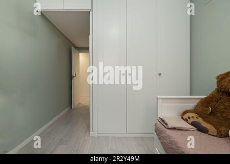 Una camera da letto con un letto piccolo e un armadio a muro con porte bianche con maniglie circolari in legno e un bagagliaio coordinato Foto Stock