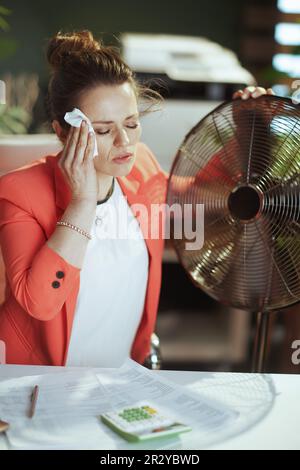 Ambiente di lavoro sostenibile. moderna donna ragioniere di 40 anni in moderno ufficio verde in giacca rossa con ventilatore elettrico e tovagliolo che soffre di estate lui Foto Stock