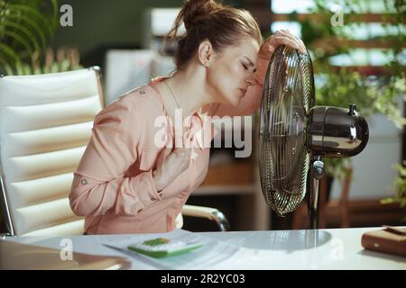 Ambiente di lavoro sostenibile. moderna donna di 40 anni al lavoro con ventilatore elettrico che soffre di calore estivo. Foto Stock