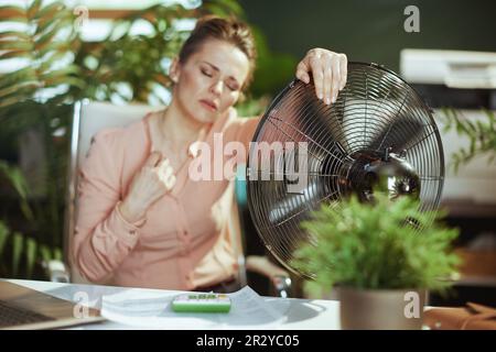 Ambiente di lavoro sostenibile. moderna donna di 40 anni lavoratore in moderno ufficio verde con ventilatore elettrico che soffre di calore estivo. Foto Stock