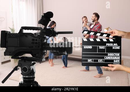 Ripresa di un filmato. Seconda macchina fotografica assistente che tiene il clapperboard vicino alla videocamera di fronte alla famiglia felice (attori) a casa (set cinematografico) Foto Stock