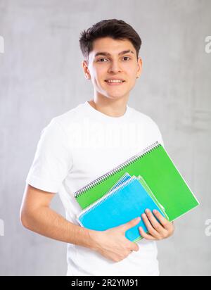 Giovane studente sorridente che si posa con i libri degli esercizi contro lo sfondo grigio ombreggiato Foto Stock