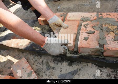 Mani in muratore che posano il primo corso di mattoni sulla parete di fondazione della casa con barriera bituminosa impermeabile. Foto Stock