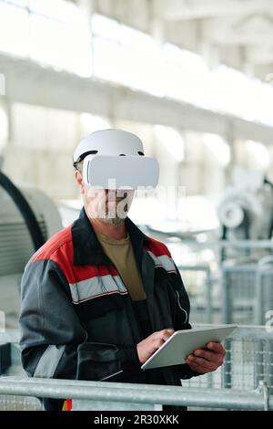 Immagine verticale di un ingegnere maturo che utilizza un tablet digitale e indossa occhiali VR per guardare il progetto online mentre lavora in fabbrica Foto Stock