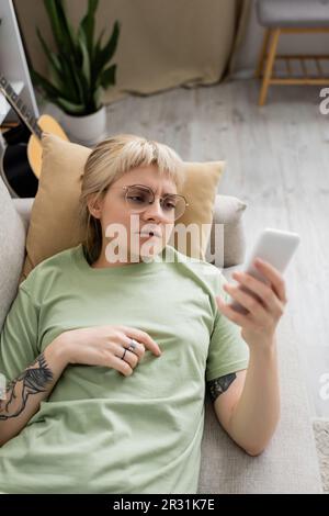 vista dall'alto della giovane donna con tatuaggio, biondo e capelli corti, scopa e occhiali con smartphone mentre si riposa su un comodo divano vicino alla chitarra Foto Stock