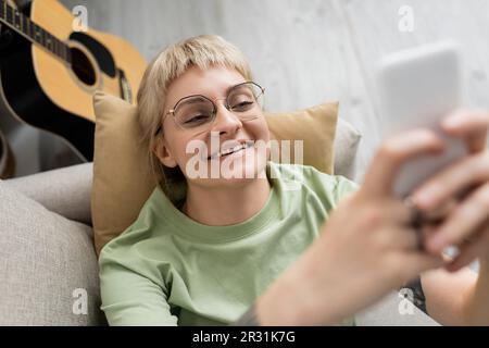 vista dall'alto di una giovane donna felice con capelli biondi e corti, scoppi e occhiali che testano sullo smartphone mentre si riposano sul comodo divano vicino a guit Foto Stock