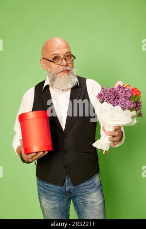 Ritratto di uomo maturo, bearato, calvo in abiti e bicchieri eleganti, in piedi con fiori contro sfondo verde studio Foto Stock