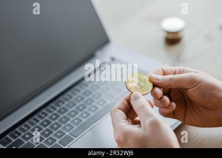 KIEV, UCRAINA - 18 OTTOBRE 2022: Vista ritagliata di un ambizioso uomo d'affari in possesso di bitcoin dorato vicino al computer portatile con schermo vuoto sulla scrivania dell'ufficio, sfocato b Foto Stock