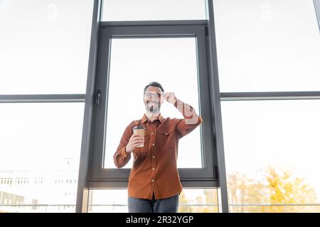 vista dal basso angolo di un allegro uomo d'affari in camicia alla moda in piedi con caffè per andare vicino a grandi finestre da ufficio, regolazione degli occhiali e Foto Stock