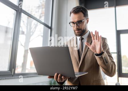 serio uomo d'affari bearded in occhiali alla moda, beige blazer e cravatta tenendo il laptop e la mano ondeggiante mentre si tiene la videoconferenza vicino vano sfocato Foto Stock