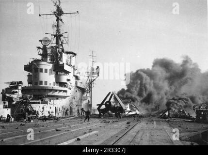 Vigili del fuoco occupati a bordo di HMS FORMIDABILE dopo che un aereo suicida giapponese si era schiantato sul ponte di volo mentre stava operando al largo delle isole Sakishima a sostegno degli sbarchi di Okinawa. Un Corsair Chance-Vought con le sue ali piegate può essere visto accanto all'isola malamente bruciata, mentre il fumo nero fuoriesce dal relitto che brucia all'estremità del ponte di volo. Foto Stock