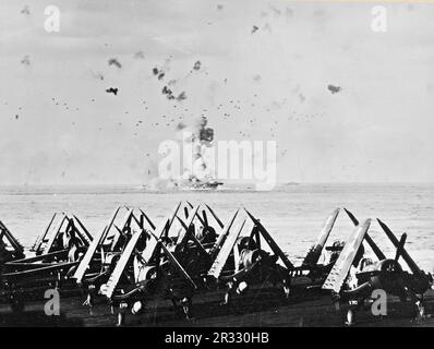 Gli Stati Uniti Portaerei Navy USS Enterprise (CV-6) in fiamme dopo che la nave è stata colpita da un kamikaze al largo di Kyushu il 14th maggio 1945. L'elevatore anteriore, che pesa molte tonnellate, può essere visto gettato in su dall'esplosione. La foto è stata scattata da USS Essex (CV-9). Vought F4U-1 i corsari della lotta contro la Boming Squadron 83 (VBF-83) sono visibili in primo piano. Foto Stock