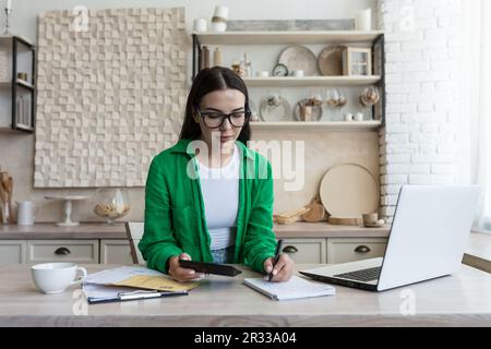 Una giovane donna lavora a casa in cucina con un computer portatile e documenti. Una casalinga si occupa di questioni finanziarie familiari e il bilancio, utilizza una calcolatrice. Foto Stock