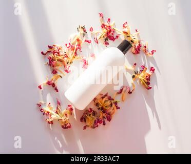 Bottiglia di plastica bianca senza marchio alla luce del sole con petali di fiori secchi intorno. Mockup per la confezione di prodotti cosmetici. Foto Stock