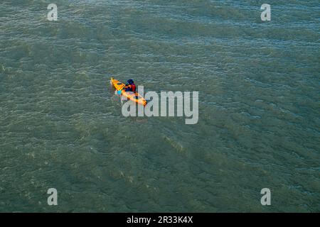 Descrizione: Una vista ad alto angolo di un individuo in un giubbotto di salvataggio che rema un vivace kayak di mare arancione attraverso l'acqua testurizzata e grigio-verdastro durante una o Foto Stock
