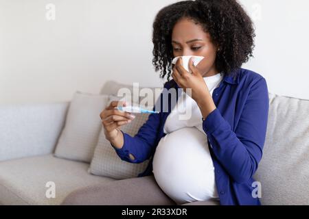 Donna nera triste riccia millenaria in abiti domestici con grande ventre soffia naso in tovagliolo Foto Stock