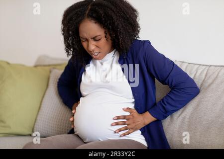 Malcontento riccio incinta donna nera millenaria in abiti domestici con grande pancia, soffre di dolore Foto Stock