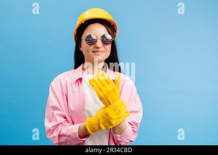 Donna con casco giallo e guanti che puntano e gesturano contro lo sfondo blu dello studio. Foto Stock