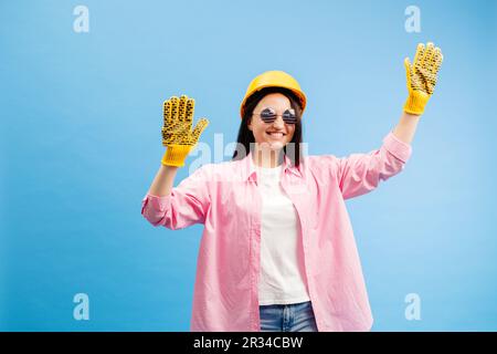 Donna con casco giallo e guanti che puntano e gesturano contro lo sfondo blu dello studio. Foto Stock