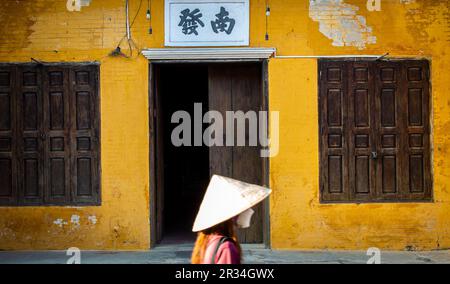 Hoian, Vietnam, 6 marzo 2023. Una donna con un cappello di riso che passa davanti a una vecchia casa con un muro giallo e una porta di legno a Hoian. Foto Stock