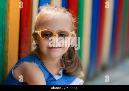 bambina con sindrome down indossare occhiali per la visione si siede vicino a una recinzione colorata in una città per bambini in estate. Foto di alta qualità Foto Stock