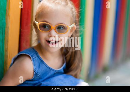 bambina con sindrome down indossare occhiali per la visione si siede vicino a una recinzione colorata in una città per bambini in estate. Foto di alta qualità Foto Stock