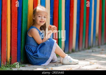 bambina con sindrome down indossare occhiali per la visione si siede vicino a una recinzione colorata in una città per bambini in estate. Foto di alta qualità Foto Stock