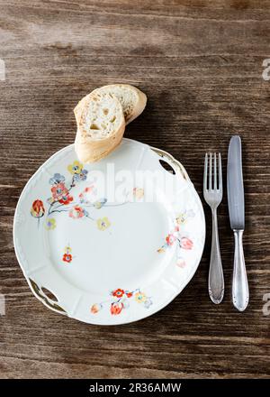 Un vecchio piatto di porcellana bianca decorato con fiori con posate in argento e due fette di baguette sul tavolo di legno Foto Stock