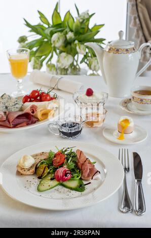 Un tavolo per la colazione con prosciutto, foglie miste, aceto balsamico, pane e burro, cetriolo, ravanelli, un uovo sodo e un piatto di formaggio Foto Stock