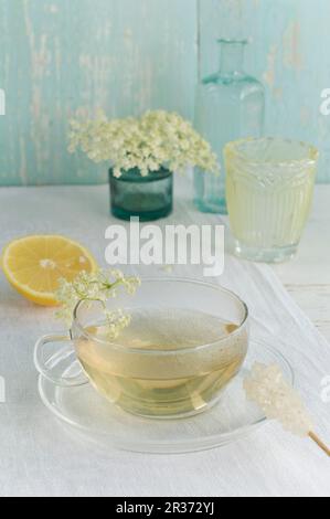Tè di fiori di sambuco in una tazza, fiori di sambuco, limone e un bastone di caramelle di roccia Foto Stock
