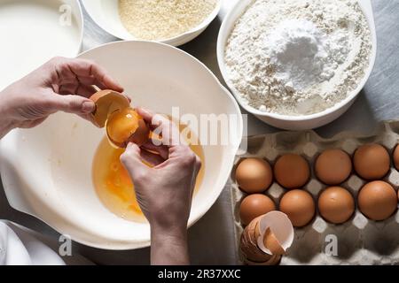 Le mani rompono le uova su una ciotola con farina di mandorle e farina normale con il lievito in polvere accanto ad essa Foto Stock