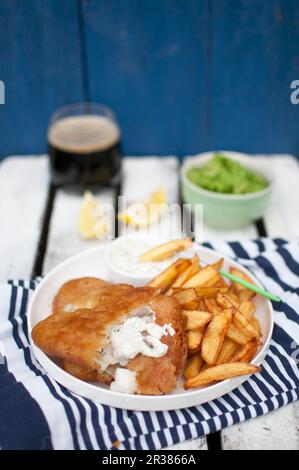 Pesce e patatine, serviti con purè di piselli verdi, pezzi di salsa tartare al limone e birra scura Foto Stock