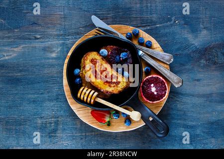 Toast alla cannella fatti in casa con miele, frutti di bosco e arancia sanguinosa in padella di ghisa d'epoca per la colazione su sfondo rustico in legno Foto Stock