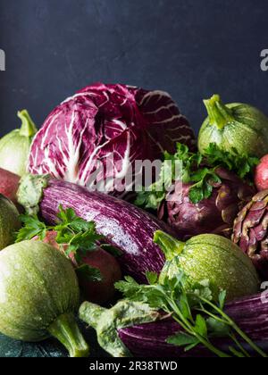 Assortimento di verdure crude verdi e viola - cicoria, patate rosse, zucchine, carciofi e melanzane - su fondo nero Foto Stock