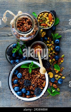 Vista dall'alto del granola al cioccolato in vaso aperto e miele servito con frutti di bosco, noci e menta fresca per una gustosa colazione su un tavolo di legno di rustin Foto Stock