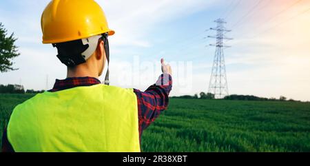 Lavoratore di costruzione che indica al pilone di elettricità. Elettricista che indossa elmetto giallo e corpetto fluorescente sul campo, zona di lavoro. OSH. Foto Stock