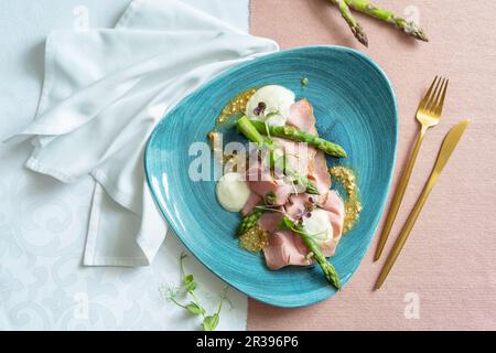 Vista dall'alto di un antipasto con asparagi, taleggio, arachidi e prosciutto al rosmarino su un piatto blu. Panno bianco e rosa, tovagliolo bianco e posate dorate. Foto Stock