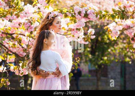 Madre alla moda con la figlia a passeggio tra sakura tree. Molla di moda femminile. Foto Stock