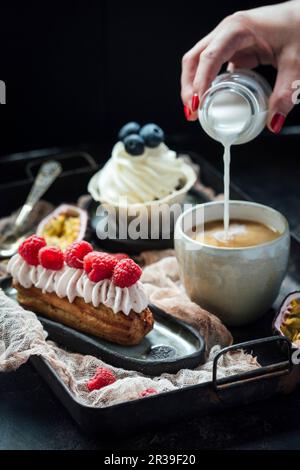 Eclair con bacche e panna, e cupcake alla vaniglia, versando il latte in caffè Foto Stock