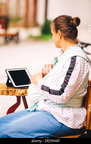 Giovane madre con bambino piccolo in sling seduto al bar all'aperto e di lavoro utilizzando un tablet Foto Stock