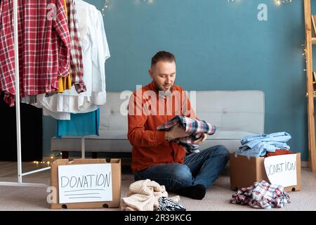 Uomo che piega vecchi vestiti in una scatola di donazione seduto sul pavimento a casa. Beneficenza e donazione Foto Stock