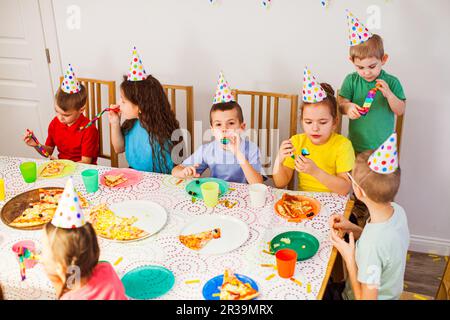 I bambini adorabili indossano cappelli da compleanno e si divertono insieme. I bambini mangiano la pizza gustosa a festa Foto Stock