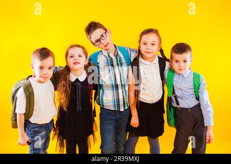 Gruppo di bambini felici in uniforme con borse da scuola abbracciando e guardando alla fotocamera su sfondo giallo Foto Stock