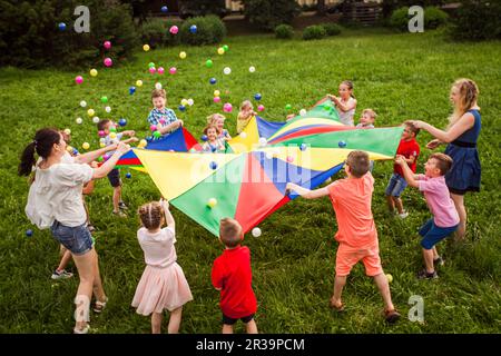 I bambini felici che sventolano il paracadute dell'arcobaleno pieno delle sfere Foto Stock