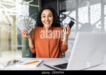 Donna ispanica di successo che guarda la macchina fotografica e sorridente, donna d'affari che tiene la carta di credito della banca e i dollari del denaro contante, ritratto della donna di successo circa, ufficio di lavoro con il laptop Foto Stock