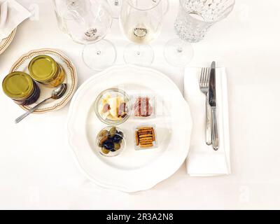 Piatto in porcellana bianca con deliziosi spuntini. Olive, formaggio, mandorle, prosciutto in coppe di vetro separate. Bicchieri di champagne vuoti. Vasetti di conservazione su p Foto Stock