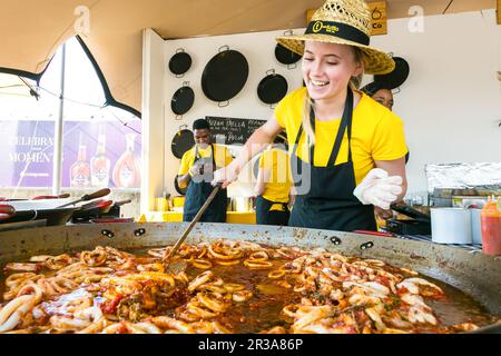 Cuoco caucasico femmina che fa e serve Paella Seafood piatti da asporto presso la bancarella del venditore del festival alimentare Foto Stock