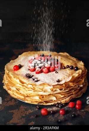 Frittelle russe con bacche cosparse di zucchero in polvere di fronte a fondo scuro. Settimana dei pancake Foto Stock