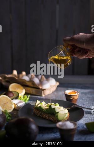 L'olio d'oliva viene versato da una brocca di vetro sul pane all'avocado Foto Stock