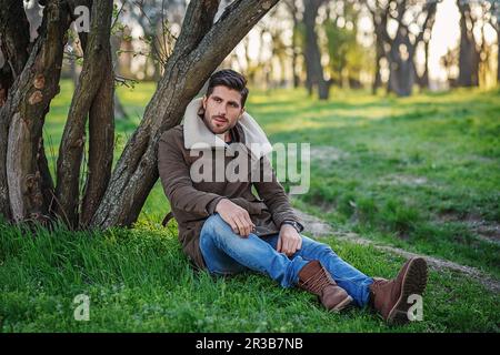 Ritratto di giovane attraente trendy seduto su erba verde in un parco al tramonto Foto Stock